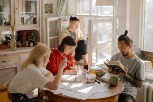 4 mensen zitten samen rond een tafel aan het raam. 2 personen lezen een boek en 2 anderen schrijven iets neer op papier.